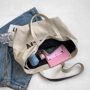 Beige letter printed canvas tote bag with large capacity, displayed open with a pink tablet, umbrella, and water bottle inside, placed on a blue denim jacket.