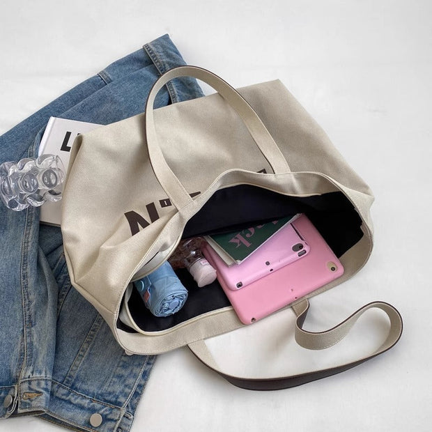 Beige letter printed canvas tote bag with large capacity, displayed open with a pink tablet, umbrella, and water bottle inside, placed on a blue denim jacket.