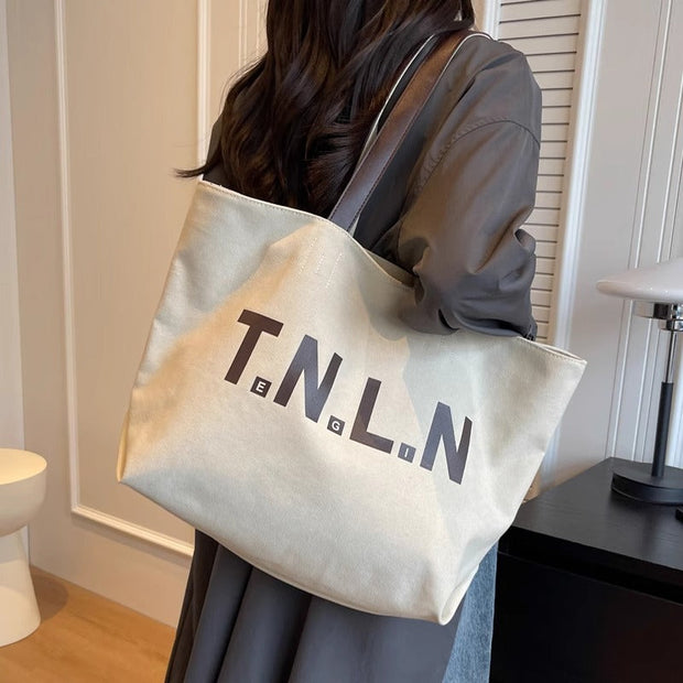 Woman carrying a large off-white canvas letter printed tote bag with black straps over shoulder in indoor setting